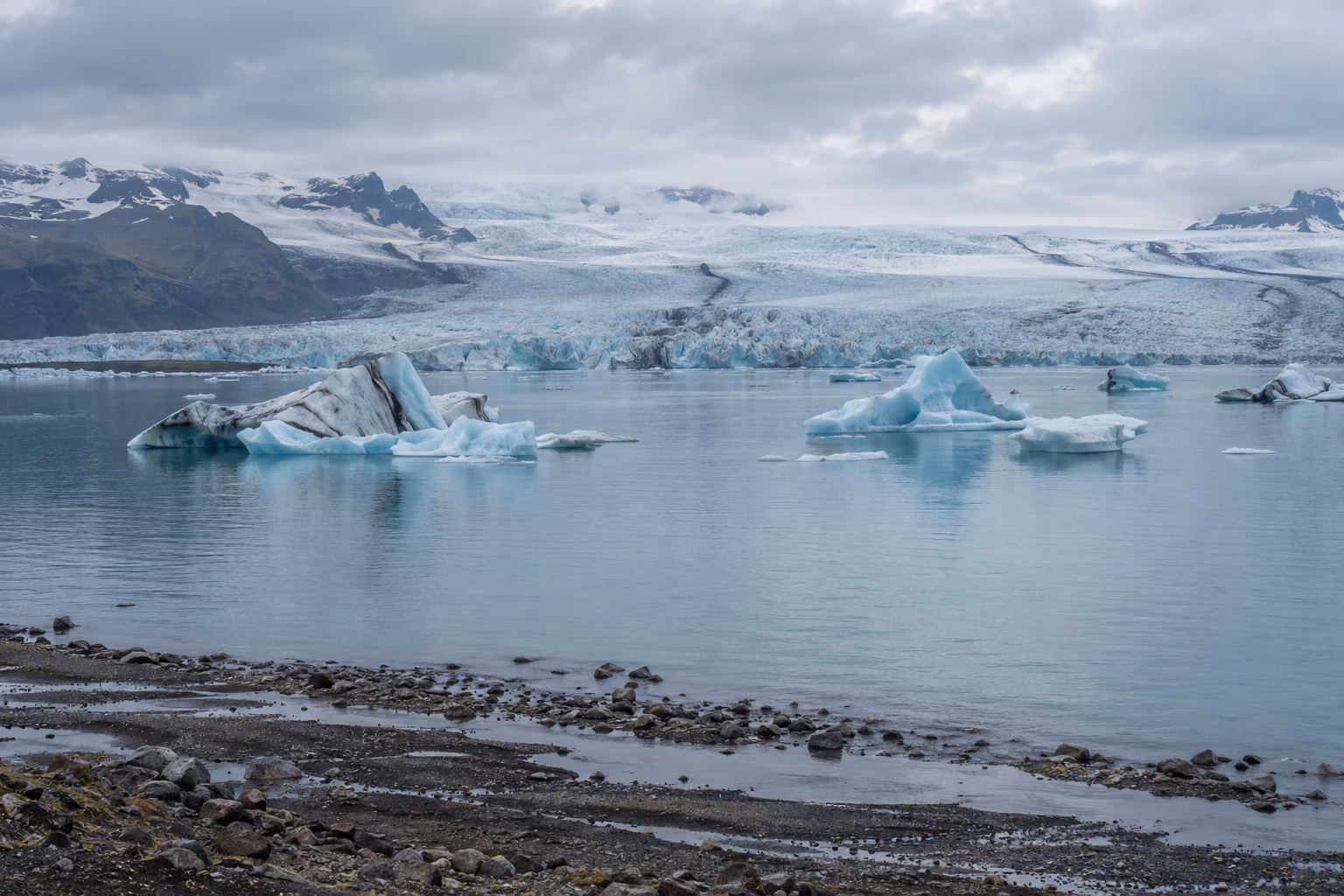 Recreación editorial de una laguna glaciar inspirada en Jökulsárlón, con icebergs, un frente de hielo y una orilla de sedimentos oscuros.