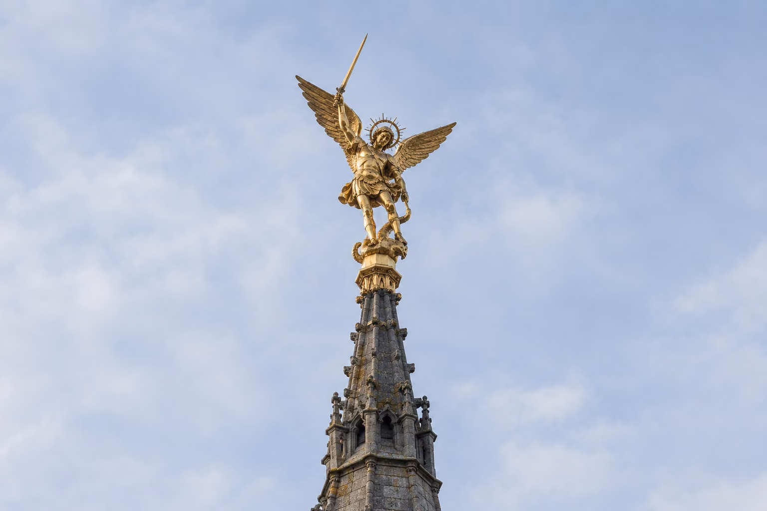 Recreación editorial de la estatua dorada de san Miguel sobre la torre de la abadía del Mont-Saint-Michel, con cielo claro.