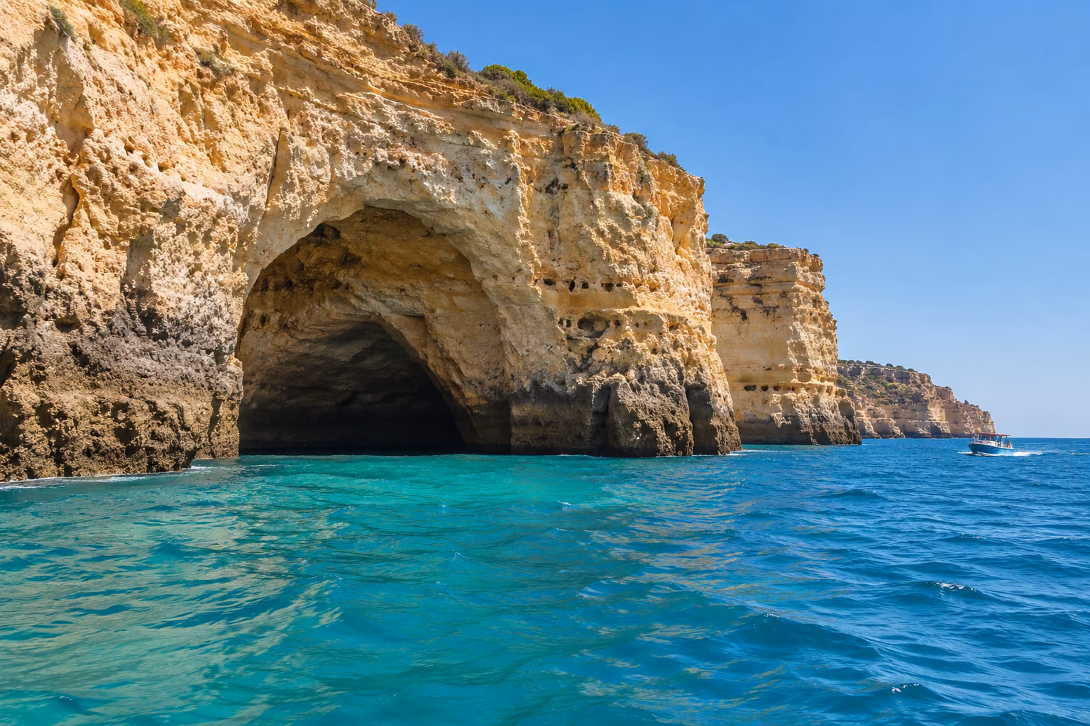 Vista exterior de una cueva marina del Algarve con un barco de excursión a distancia, sobre el agua.