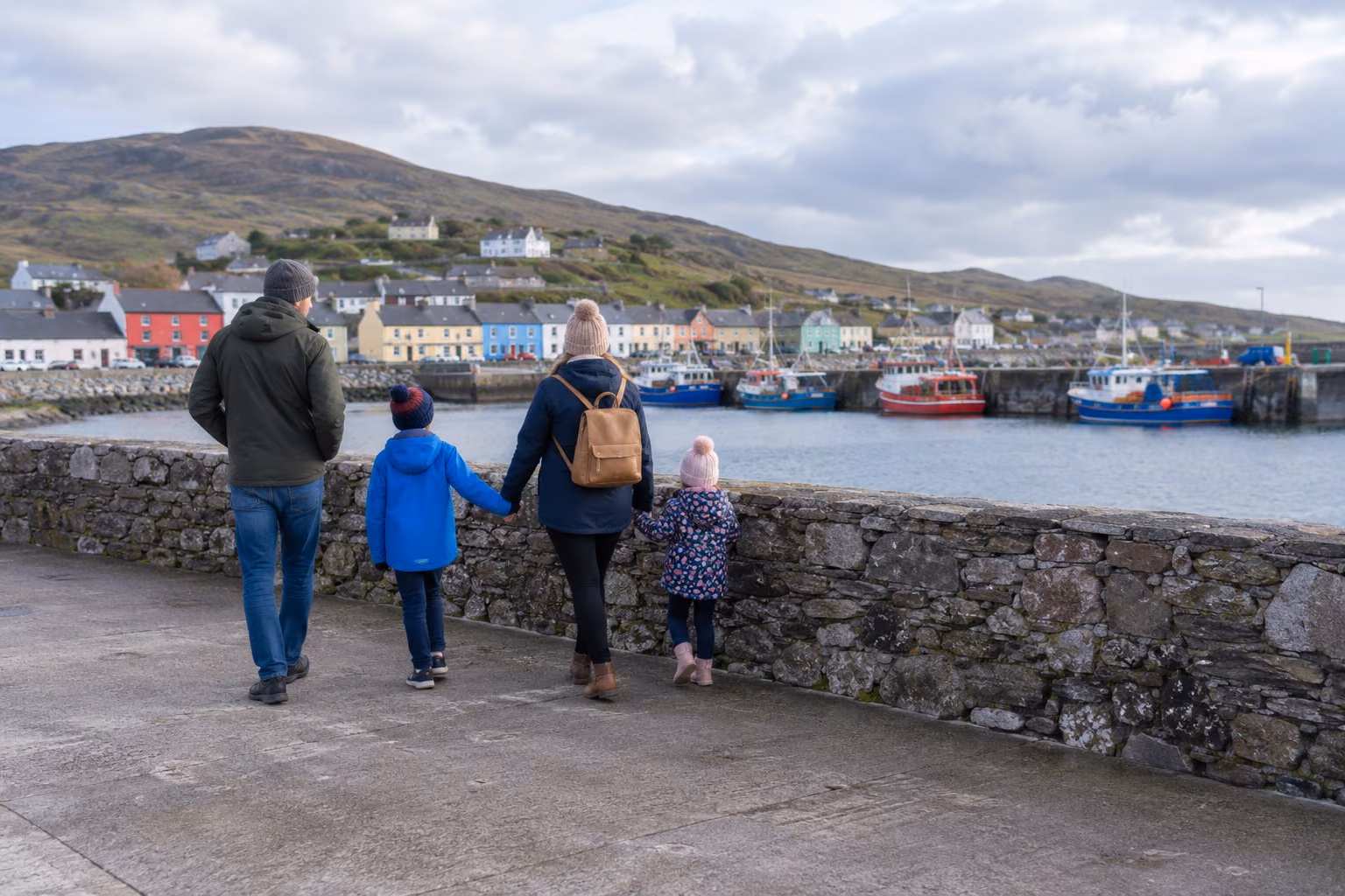 Familia pasea por el puerto de Portmagee, tras un muro de piedra y con barcos al fondo; recreación editorial, no fotografía documental.