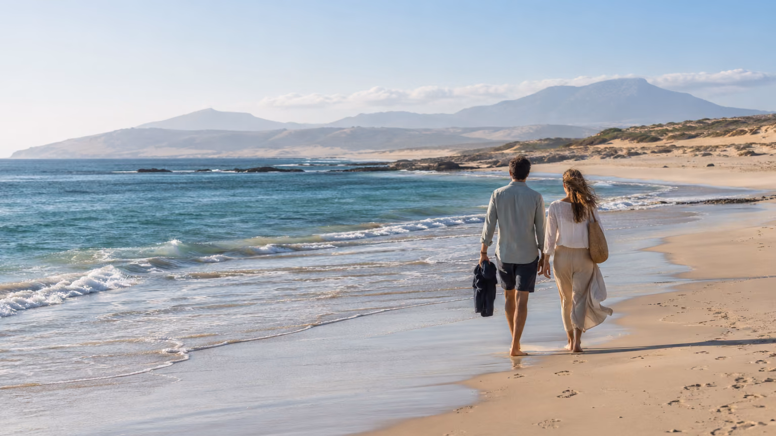 Dos viajeros caminan junto al mar en una costa atlántica amplia, con colinas bajas al fondo; imagen editorial generada, no es una fotografía documental de Tarifa.